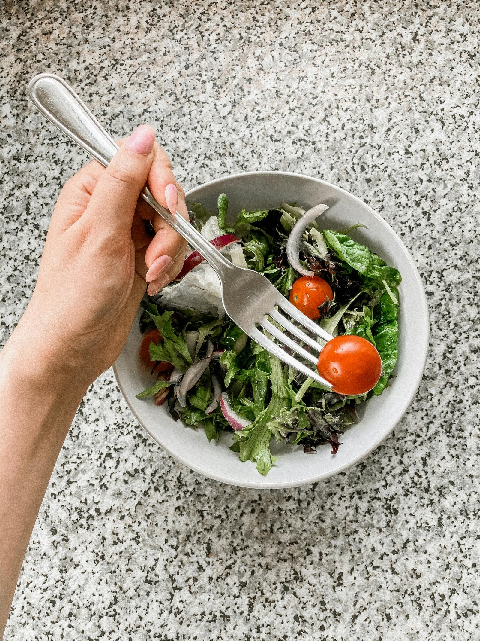 Comece Aqui A top view of a fresh, mixed green salad with cherry tomatoes in a bowl, featuring a hand holding a fork.
